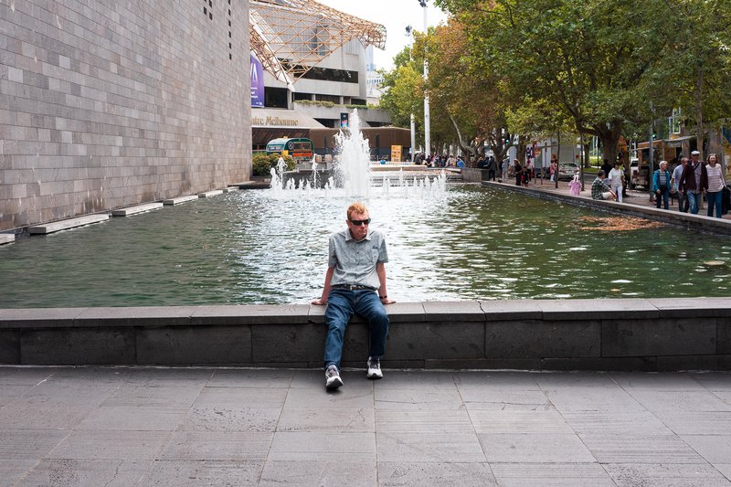 A man sits on a stone wall in front of a large fountain and a building.