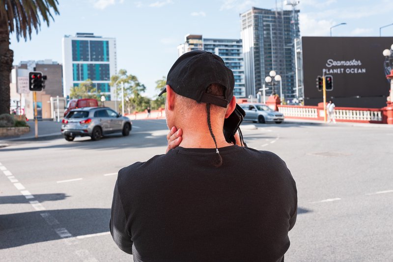 A man in a black shirt and cap stands at a crosswalk, looking away from the camera.