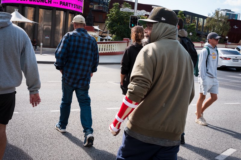 A crowd of people walking on a city street, with a man in the foreground wearing a green hoodie.