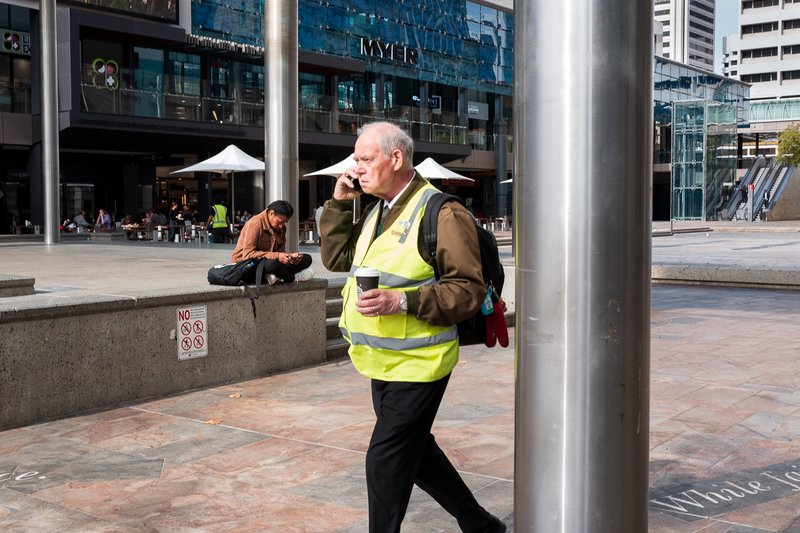 A man in a high-visibility vest walks on a sidewalk in front of a city building.