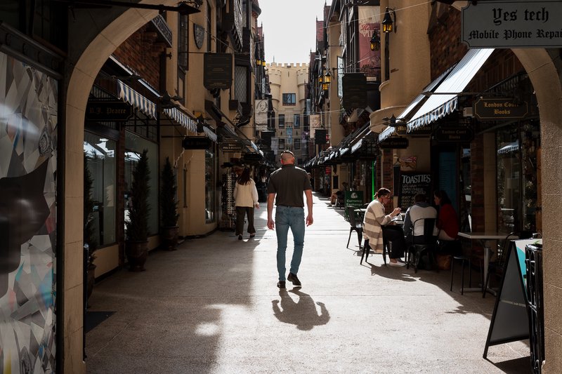 A man walks down a narrow, sunlit alleyway lined with shops and restaurants.
