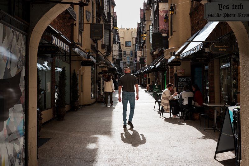 A man walks down a narrow, sunlit alleyway lined with shops and restaurants.