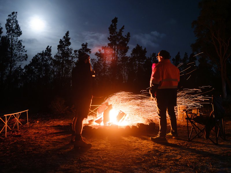 Two people stand by a fiery campfire under a bright moonlit sky.