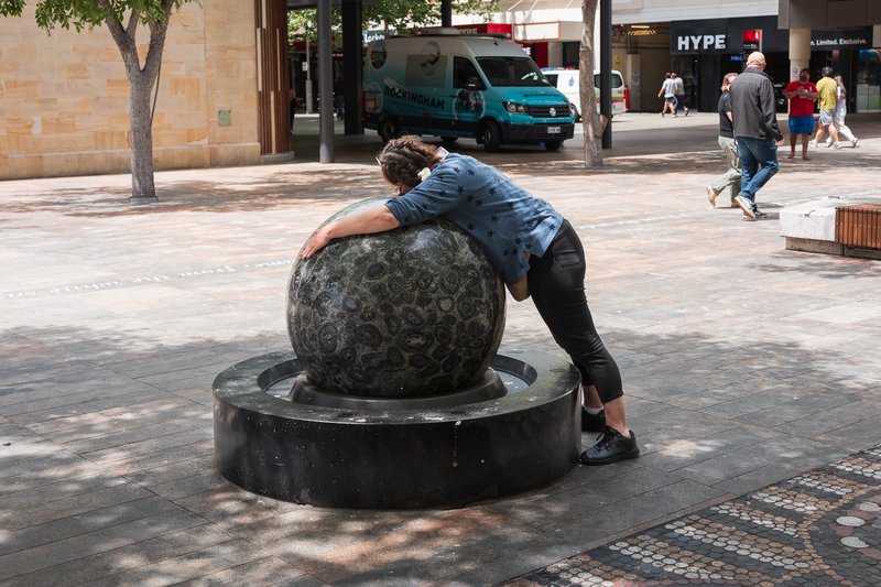 Woman leans on large spherical sculpture in bustling city square.